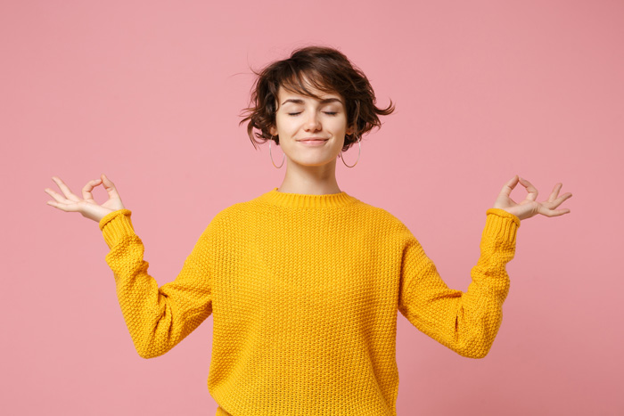 pretty young woman in yellow sweater with eyes closed and hands making meditation symbol on pink background - your recovery