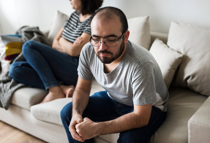 couple sitting on couch at home seem to be in an argument - woman has arms crossed and man looks sad and upset - interventionist